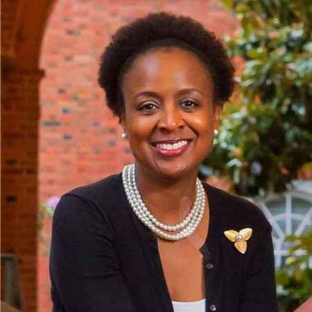 Kisha Petticolas is a Black woman with short natural hair and is smiling at the camera. She is wearing a pearl necklace and a dark cardigan. She is outside and has a brick wall and tree behind her.