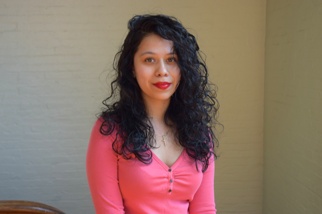 Smiling person with pink shirt, a necklace cross, and long black hair. 