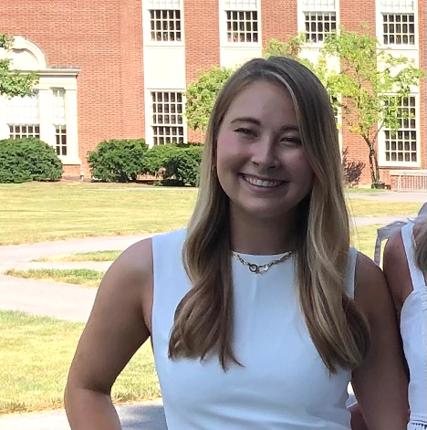 Olivia Spacassi has a light skin tone, long loose wavy blondish brown hair, has on a white sleeveless top,  and is smiling at the camera. She is outside standing a little ways away in front of a brick building.