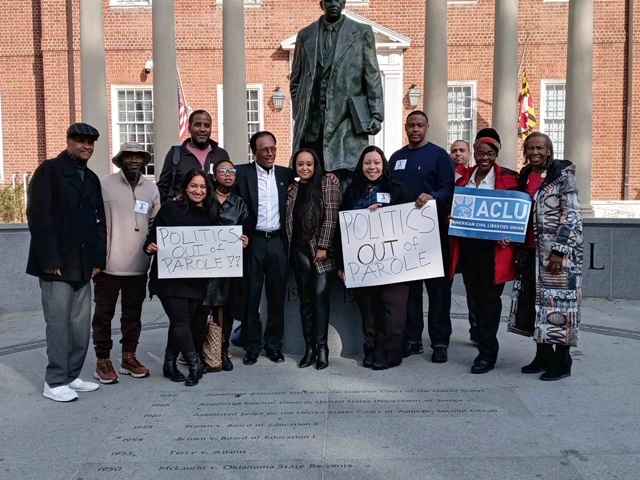 Group of advocates celebrate the veto override of parole reform in Annapolis, Maryland. They stand in front of the Thurgood Marshall statue. Some hold signs about getting politics out of parole and one person holds a sign with the ACLU-MD logo.