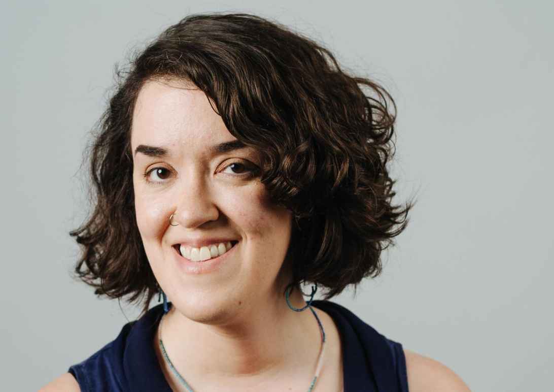 Sarah Dwyer is a white woman with chin-length dark wavy hair. She is smiling at the camera and is wearing a dark sleeveless top. The background is a gray wall.