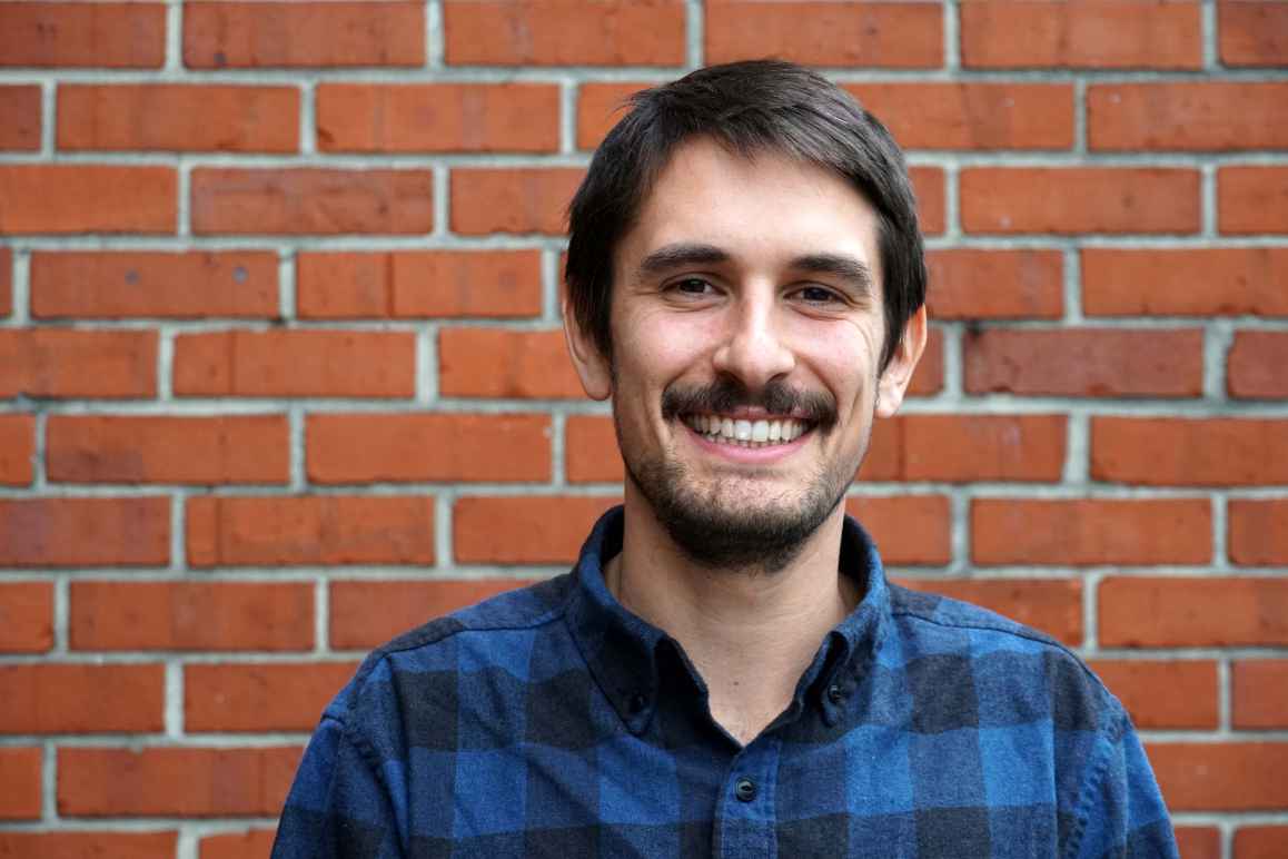 Sebastian Brown is wearing a blue and black flannel, is smiling, and is shown from the shoulders up in front of a brick wall.