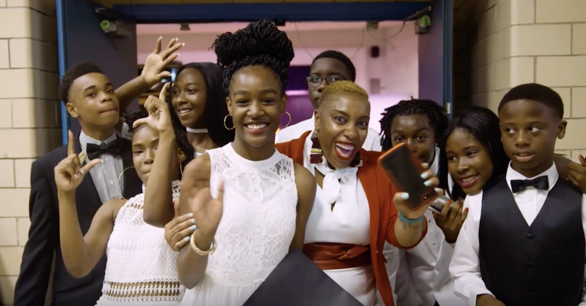 Shantay McKinily is pictured surrounded by about ten students at a formal school event. Studetns are wearing dresses and suits. Everyone is smiling.