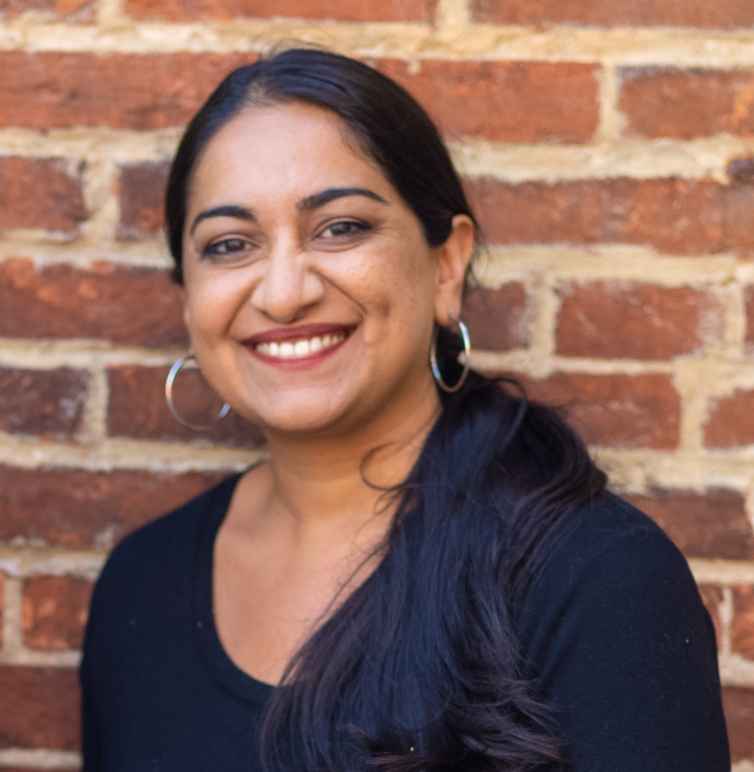 Sonia Kumar is a Middle Eastern woman with warm mediu brown skin tone. She is smiling at the camera and has long black hair pulled back in a low pony tail. She is wearing hoop earrings. She is standing in front of brick wall.