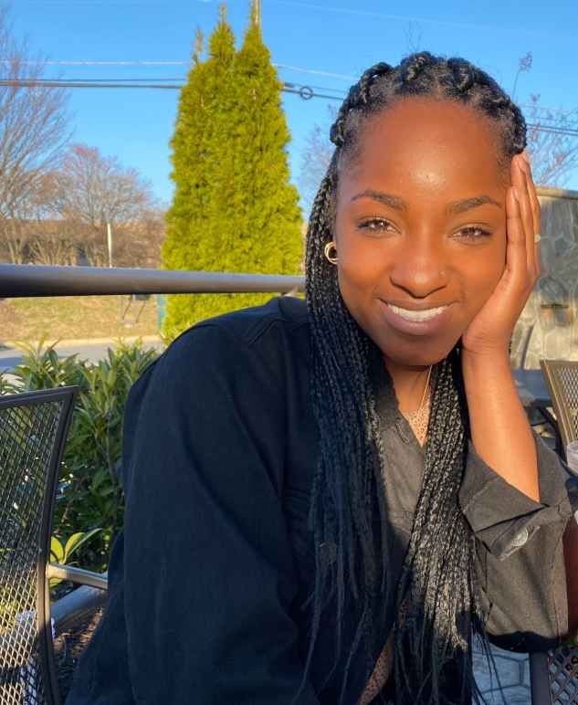 Sydney Moore is a Black woman picture with long braids. She is smailing and looking at the camera with her left elbow on a table and hand on the side of her face. She is smiling and is wearing a dark jacket and long-sleeve shirt. She is sitting outside.