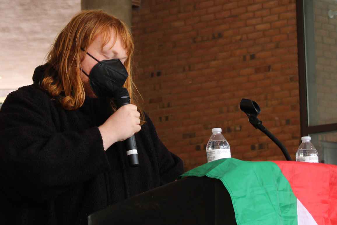 Chessie Jackson has shoulder-length red hair, is wearing a mask, and is speaking into a microphone. There is a Palestinian flag draped next to her.