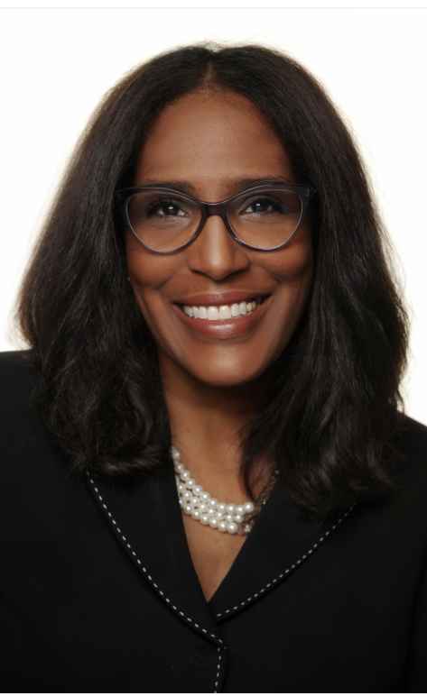 Shawn Essien is a Black woman who is smiling at the camera, has medium-length hair with light waves, rich warm Brown skin tone, is wearing glasses, a pearl necklace, and a black suit jacket.