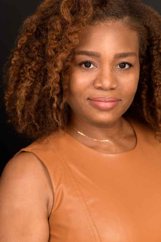 Veronica Dunlap is a Black woman with shoulder length curly hair and warm highlights. She is looking at the camera. she is wearing a sleeveless caramel-colored shirt.