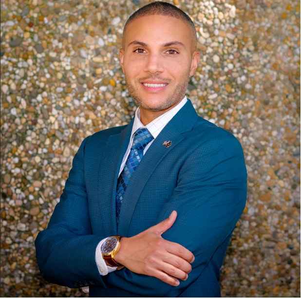 Jordan Howlette is a man with a latte skin tone, very short hair, is wearing a blue suit jacket, white button-up shirt, blue tie and has on a watch. He is smiling at the camera and standing in front of a sparkly wall.
