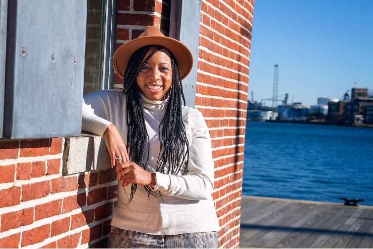 Oprah Keyes is a woman with chestnut skin tone, has long dark hair with small braided style. She is wearing a brimmed hat and white long sleep shirt. She is standing outside leaning on a brick wall with a dock and body of water in the background.