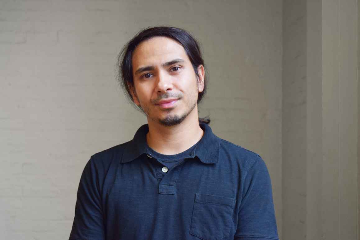 Frank Patinella is a multiracial Thai American. He is standing in front of a light wall, has dark hair pulled back, and is wearing a navy blue collared cotton shirt.