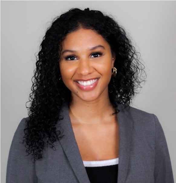 Yasmin Behram is a woman with a hazelnut skin tone, has medium-long curly dark hair, is wearing a gray blazer and black top with white piping. She is wearing rosy lipstick and is smiling at the camera. The background is a solid gray.