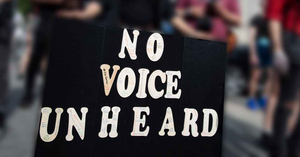 A person's hands are holding a large protest sign that says, "No Voice Unheard." The background is blurred out but you can see that there are people at a protest. Courtesy credit: Molly Kaplan, ACLU. 