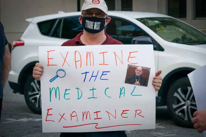 Person holds a rally sign that says, "Examine the Medical Examiner."