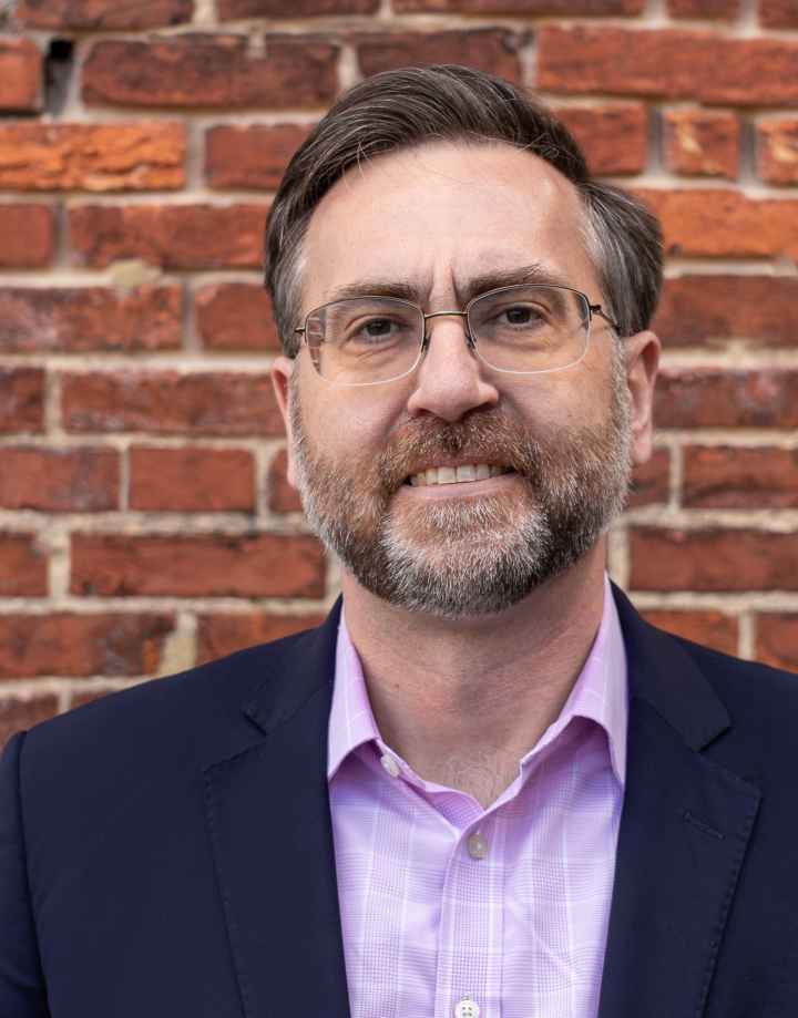 Corey Stottlemyer is standing in front of a brick wall. He is a white man with short brown and gray hair, wearing glasses and a suite.