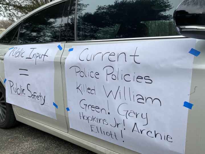 Rally signs are taped to the side of a car. They say, "Public Input = Public Safety. Current Police Practiceds Killed Willian Green, Gray Hopkins, Jr., Archie Elliot!