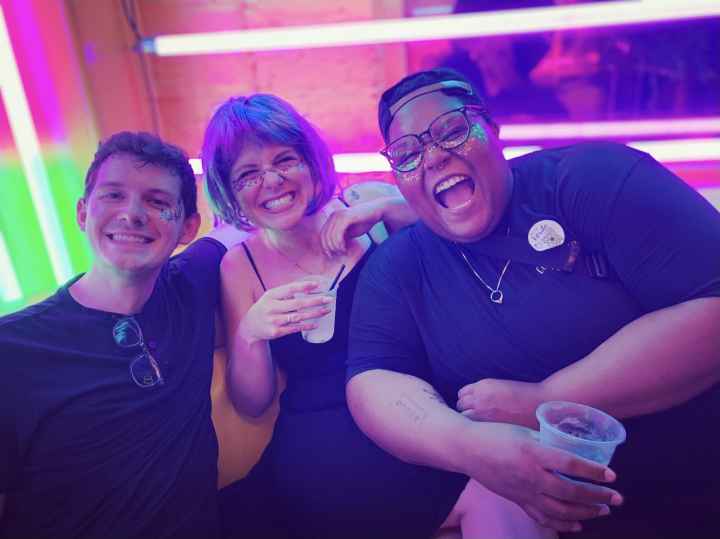 Alicia Smith on the far right is a Black, queer, non-binary person and is smiling and laughing with two friends. The background is colorful. Alicia has glitter on their face.