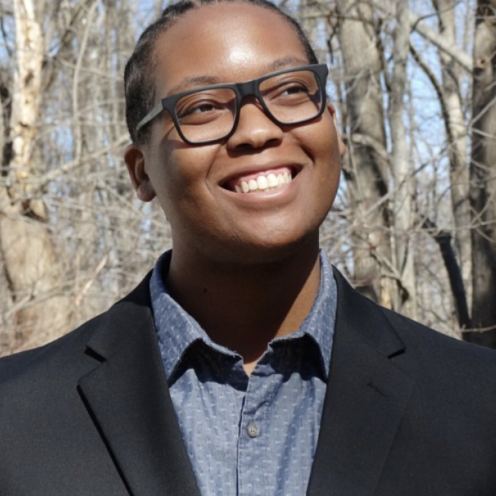 Carlos Childs is looping to the side and up and is smiling. He is a Black man and is earing dark0framed glasses. He is outside in front of trees, is wearing a black suit jacket over a blue button-up and collared shirt.