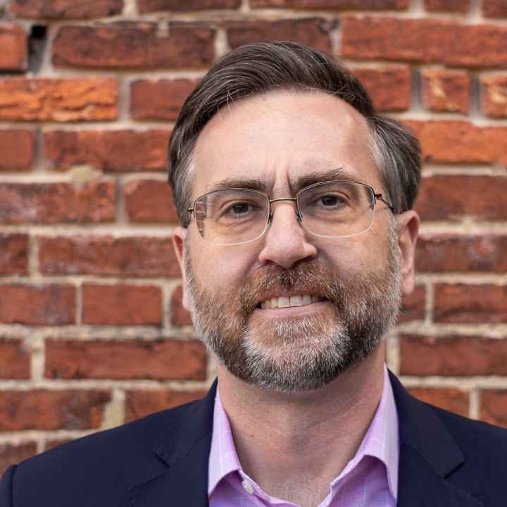 Corey Stottlemyer is standing in front of a brick wall. He is a white man with short brown and gray hair, wearing glasses and a suite.