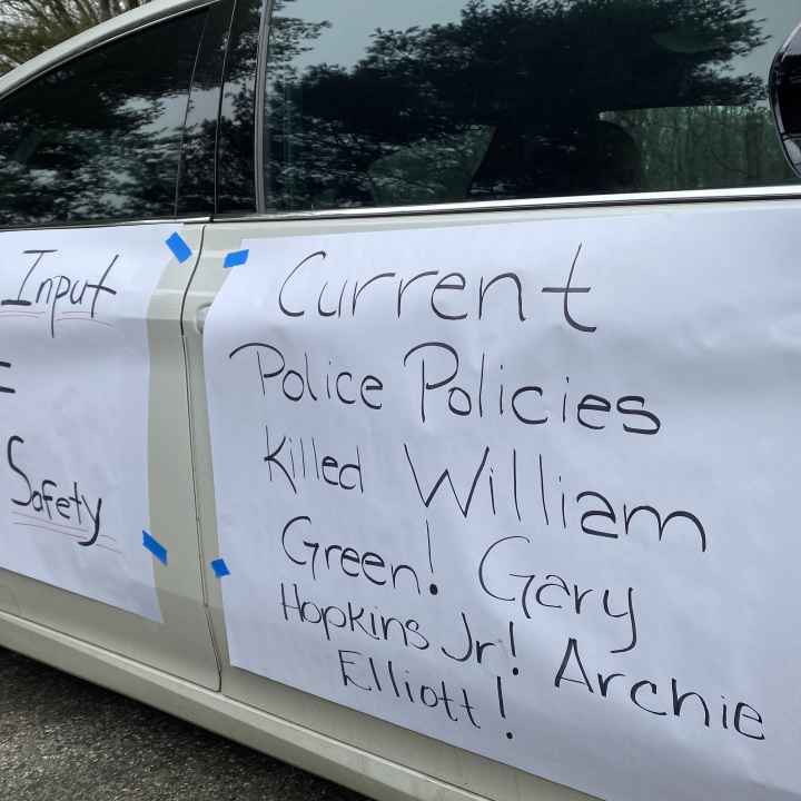 Rally signs are taped to the side of a car. They say, "Public Input = Public Safety. Current Police Practiceds Killed Willian Green, Gray Hopkins, Jr., Archie Elliot!