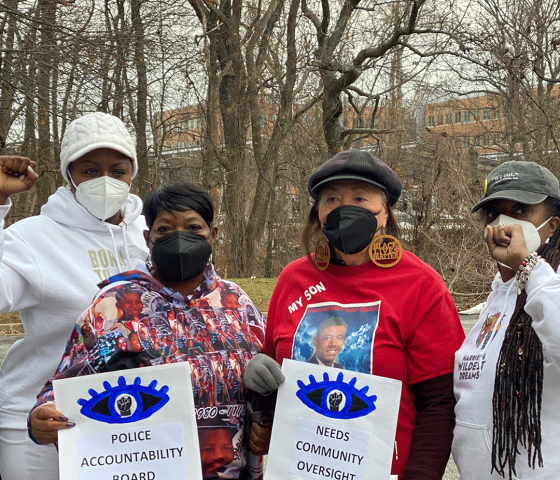 A group of Black and Residents of Color who are also advocates in Prince George's County are standing together. Two have their fists raised and two are holding signs that say "Police Accountability Board."